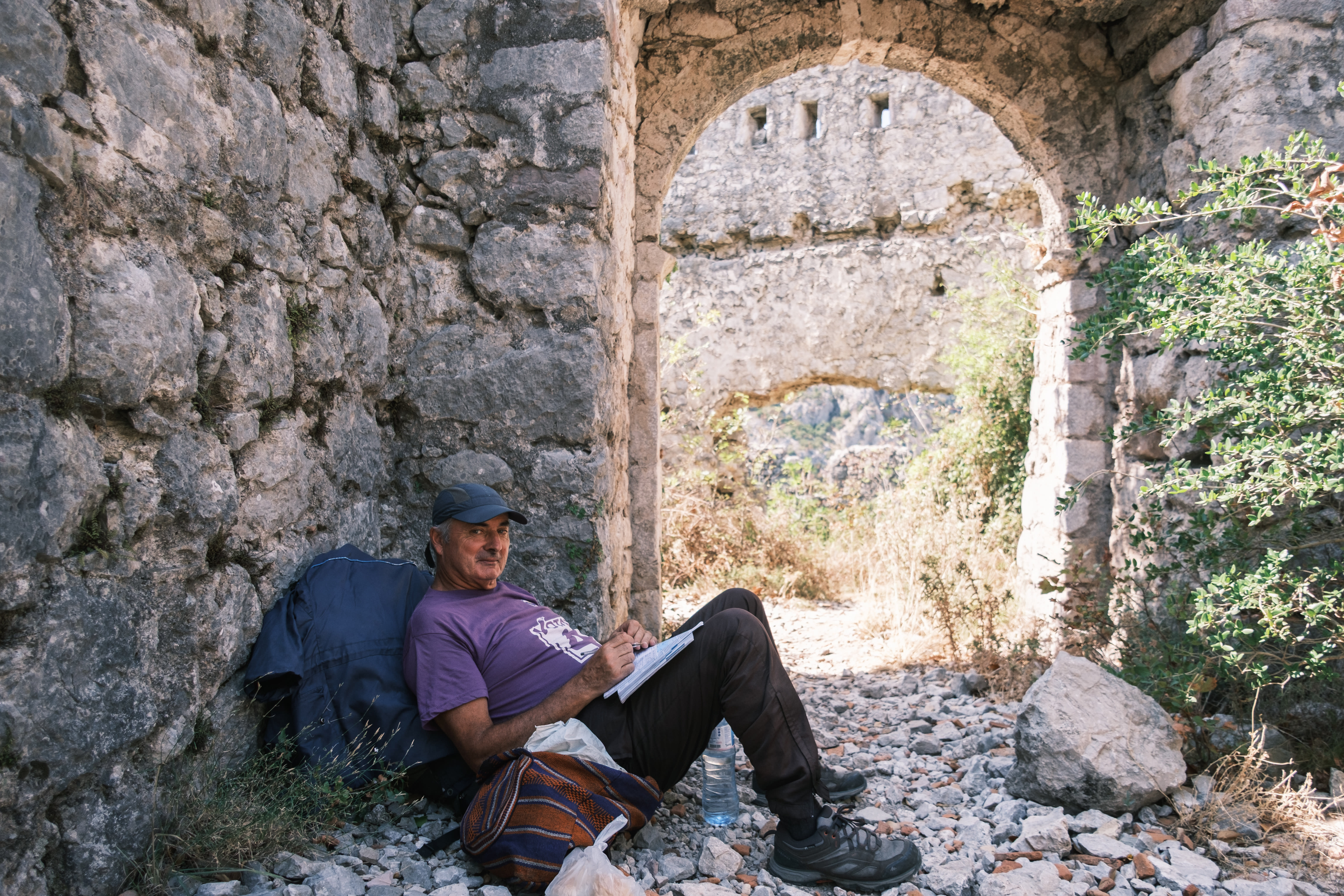 A man in a cap sketching in a notebook, leaning against a ruined stone wall beside an archway