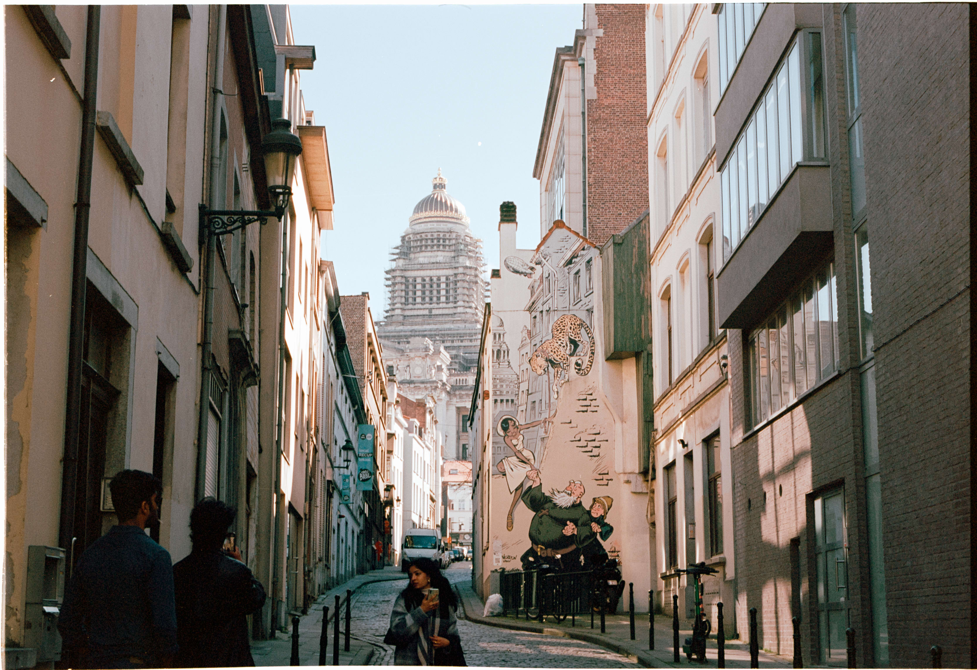 A narrow street with a comic mural, looking toward the dome of the Palace of Justice