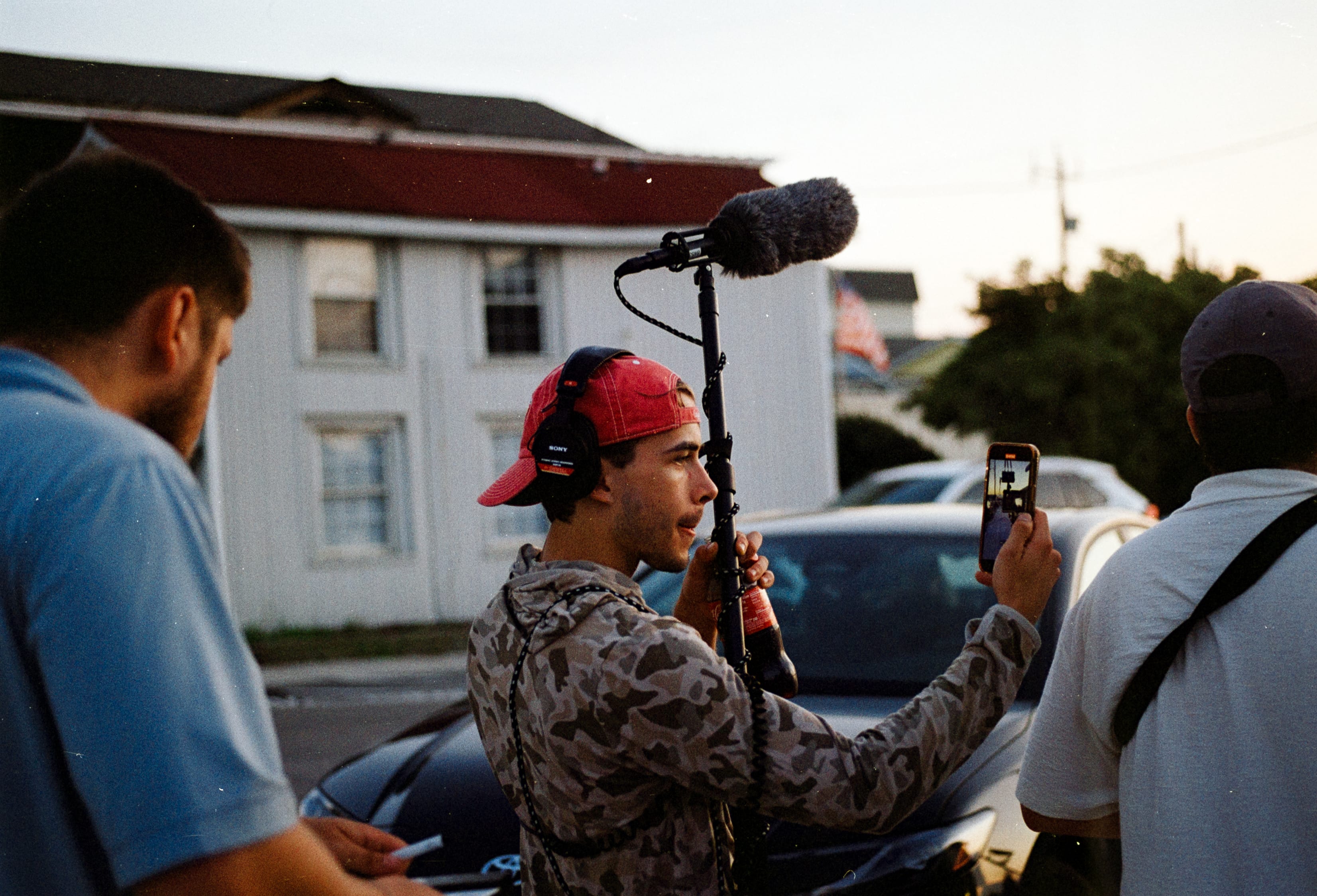 A boom operator in a red cap recording sound beside a parked car