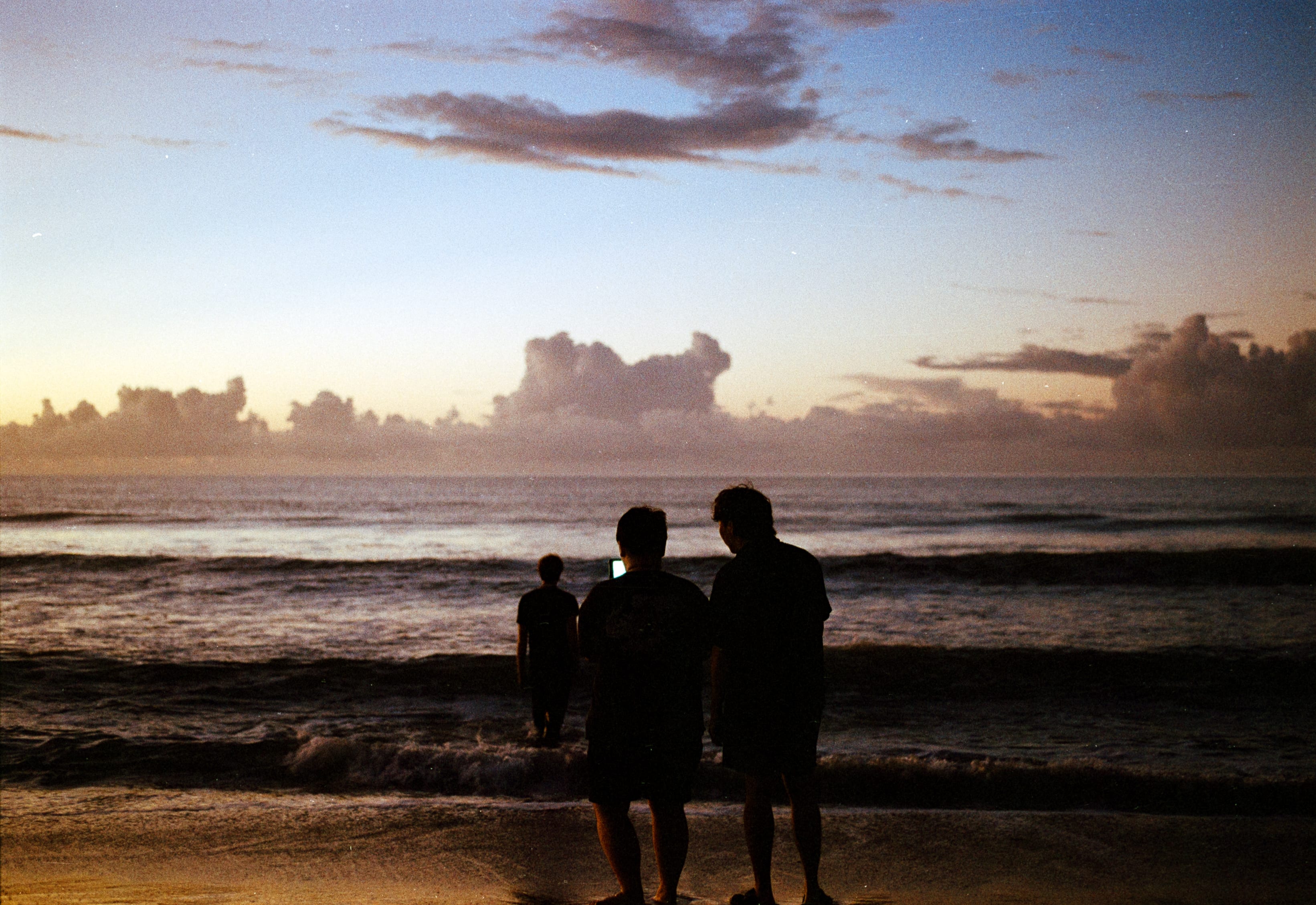Three people at the water's edge under pink clouds