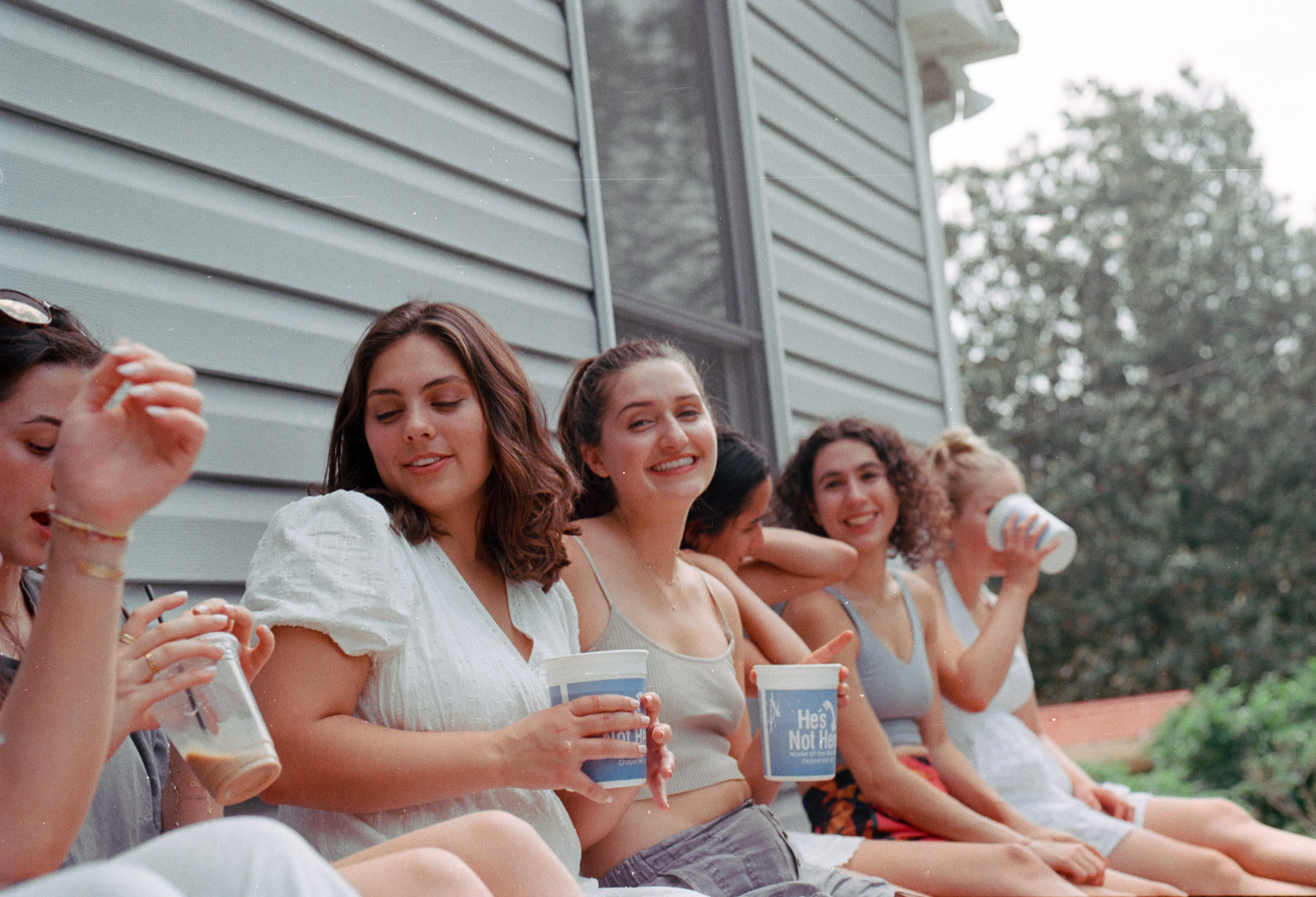 Friends sitting along the side of a house, drinking from paper cups
