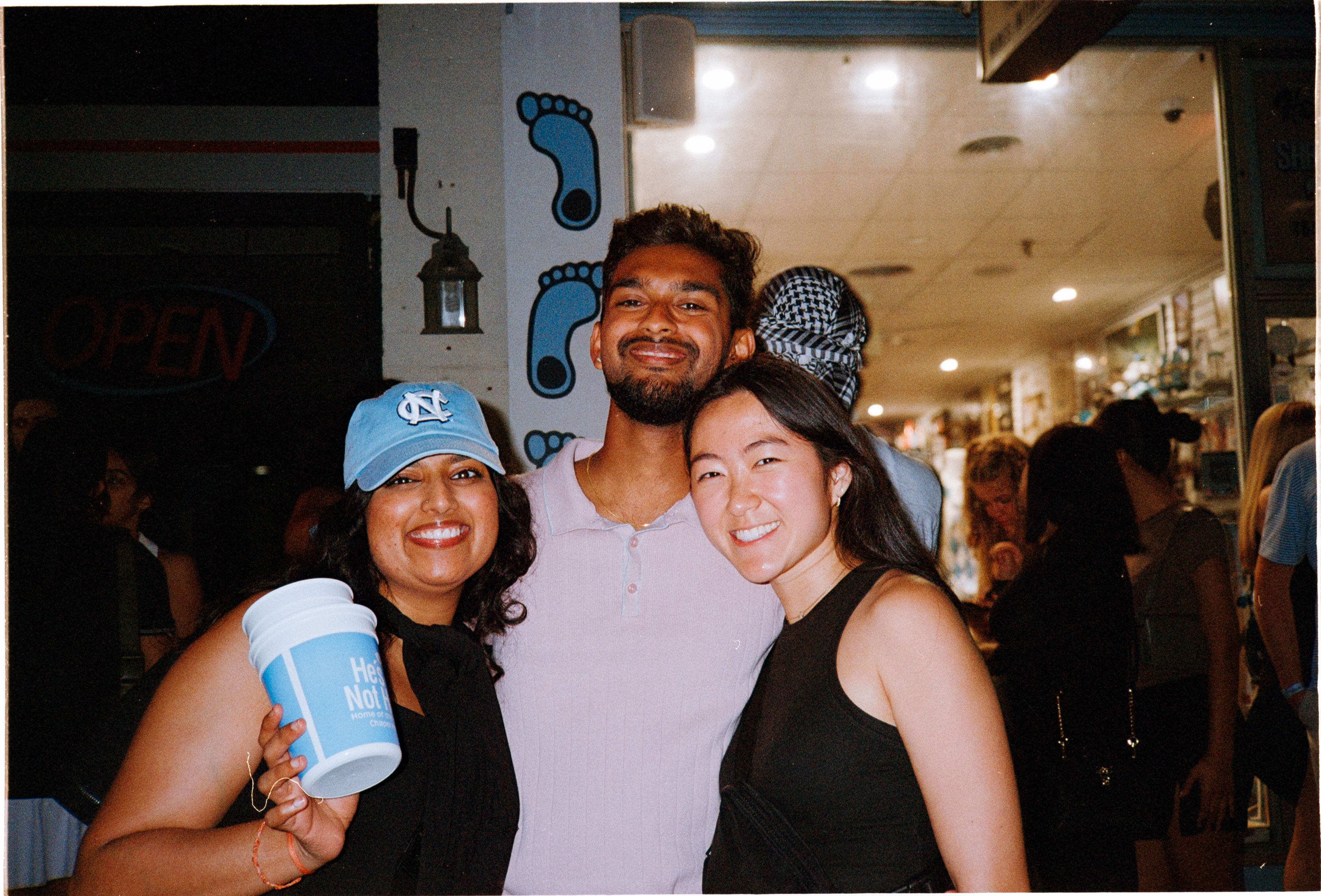 Three friends posing together at a busy spot at night, one in a Carolina cap