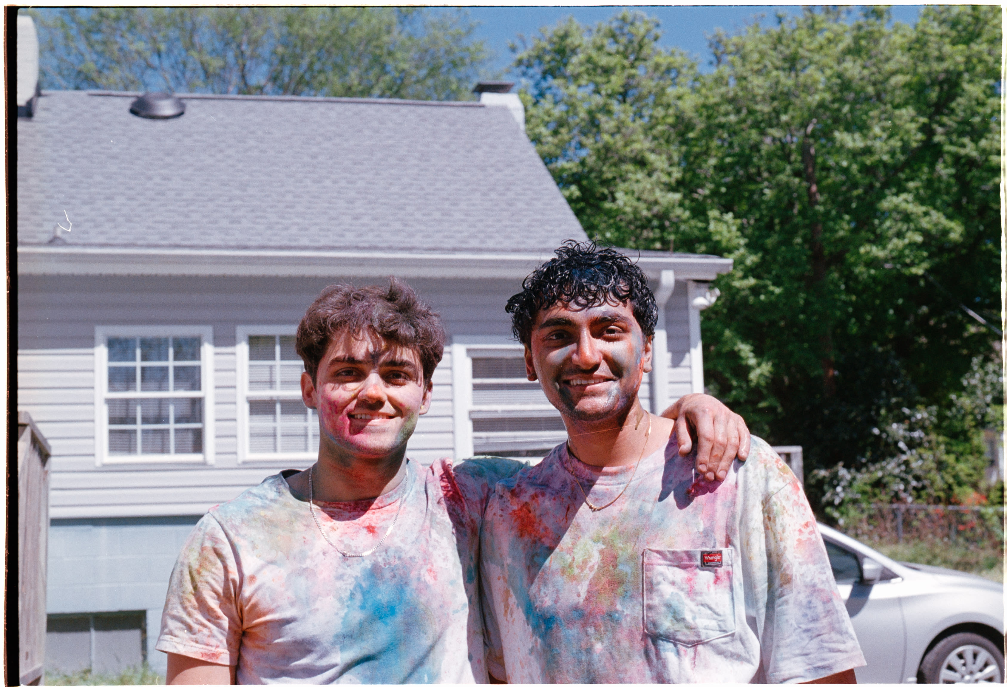 Two friends covered in colored powder, arms around each other in front of a house