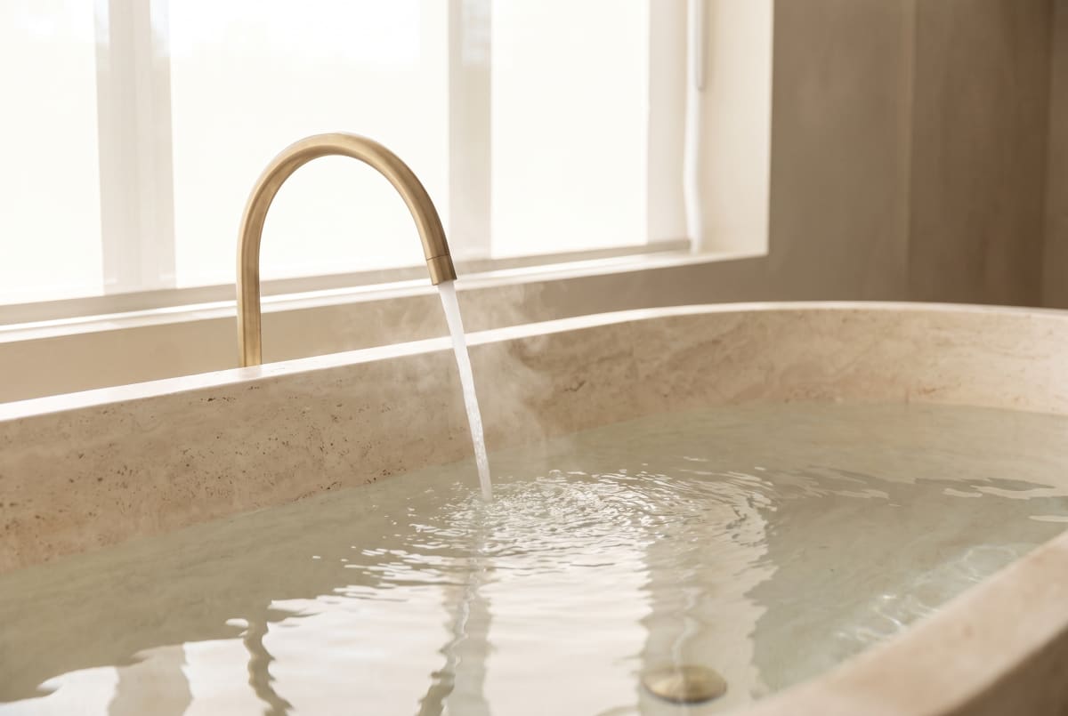Clear water filling a honed stone bath from a brushed-brass spout, soft steam.