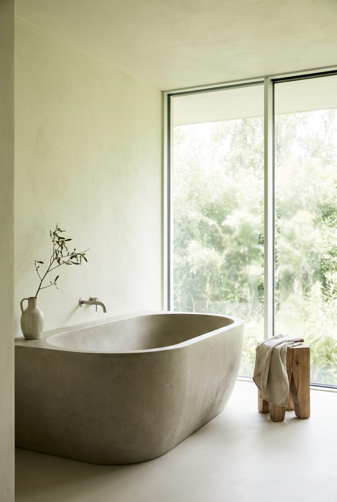 A sculptural stone bath beside floor-to-ceiling windows over green foliage.