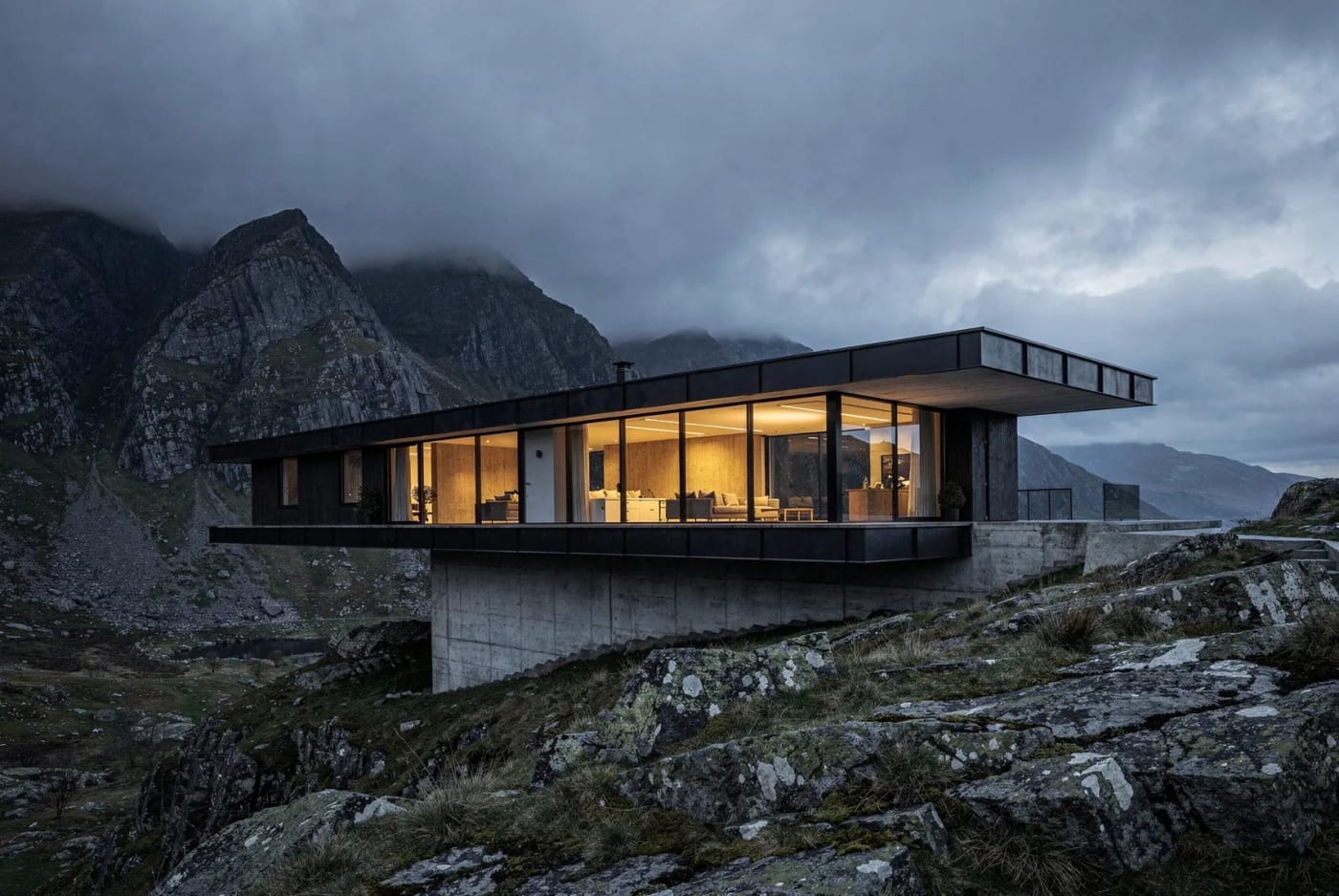 Caer at dusk — a glass and concrete house cantilevered over a Welsh mountainside, lit from within.