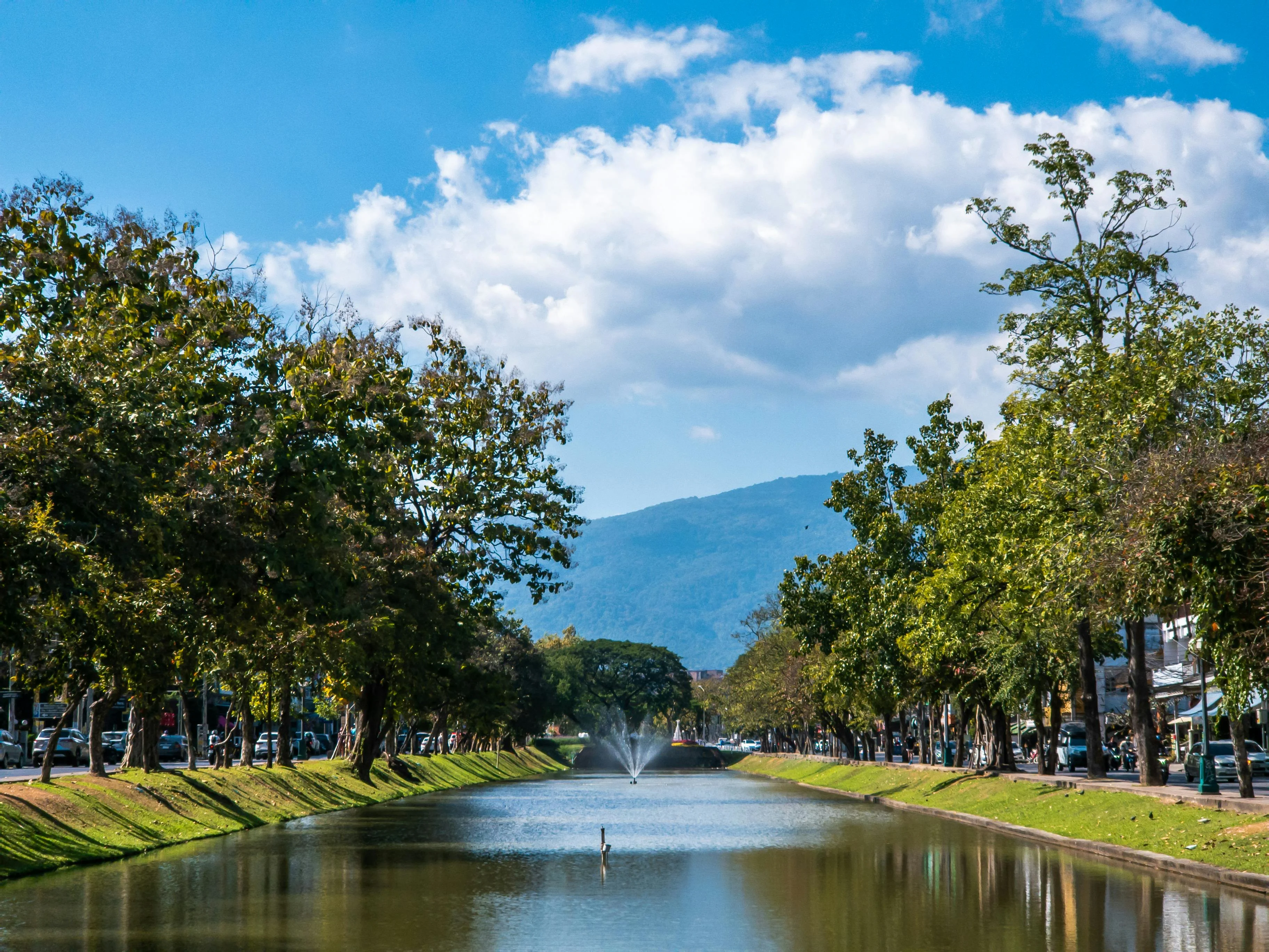 A tranquil view of a canal lined with trees and a fountain under a clear blue sky.