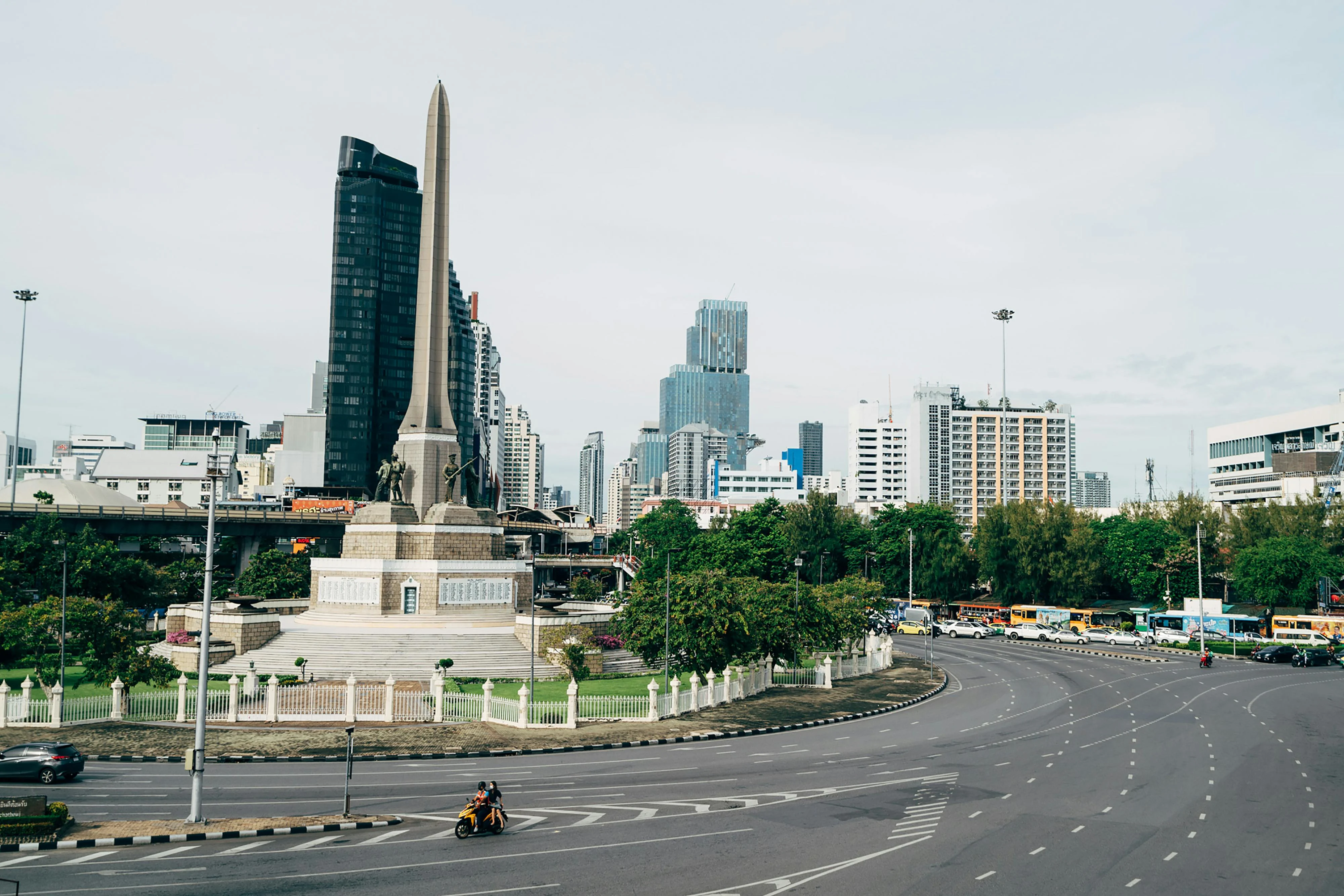 A view of the iconic Victory Monument in Bangkok, surrounded by urban skyline and traffic. More info Share