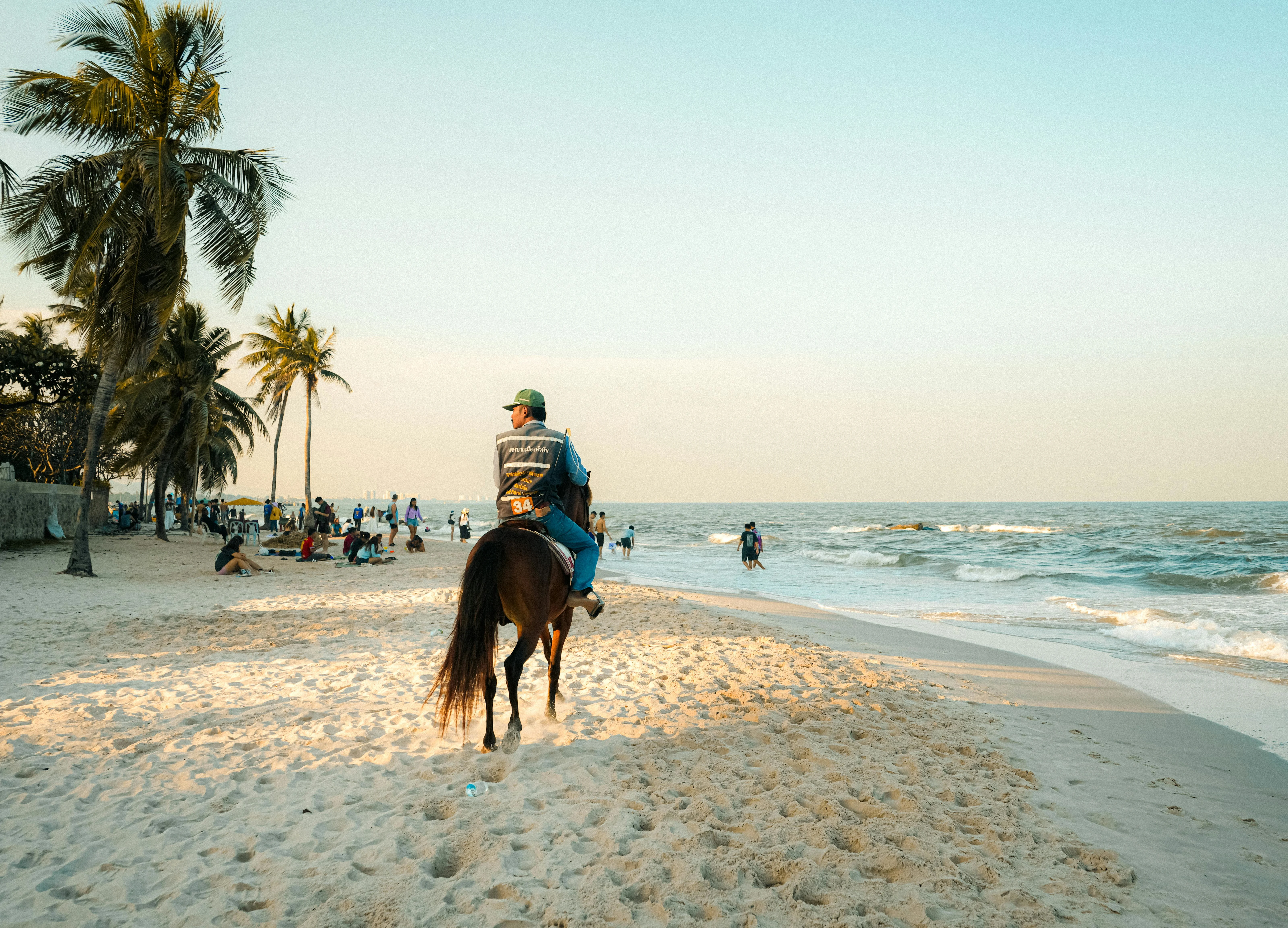 A person riding a horse on a sunny beach in Thailand, with palm trees and people enjoying the shore.
