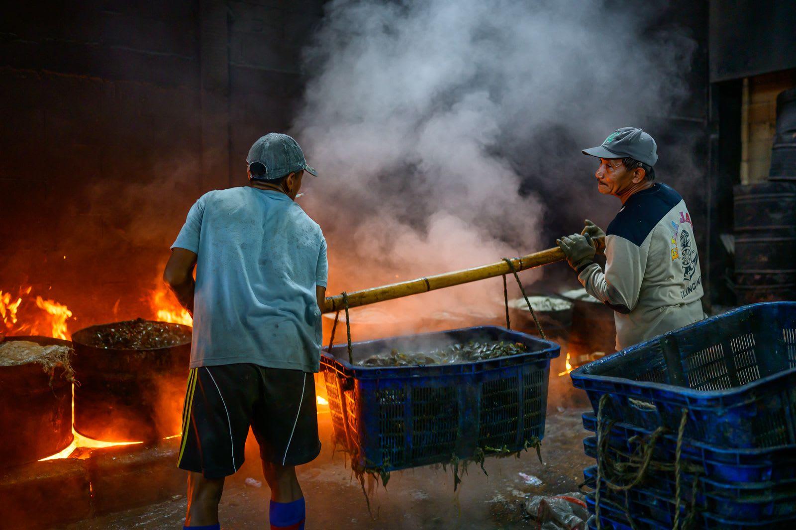 Two men carry a blue basket of steaming mussels on a bamboo pole past the cooking fires.
