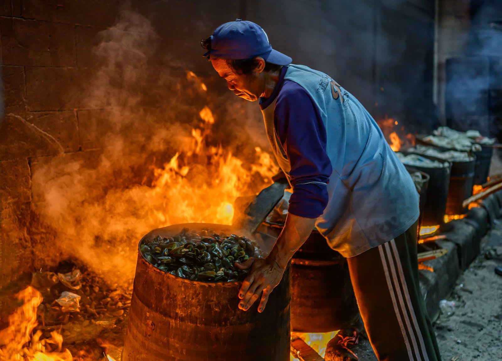 A man in a blue cap leans over a barrel of mussels cooking above open flames.