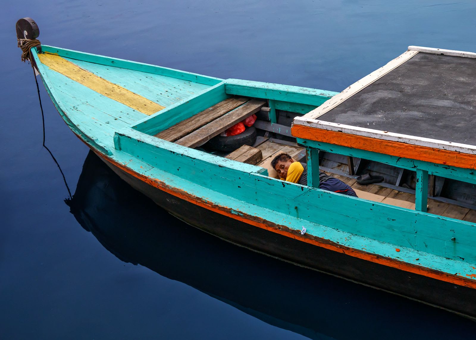 A worker rests inside a turquoise wooden boat on dark, still water.
