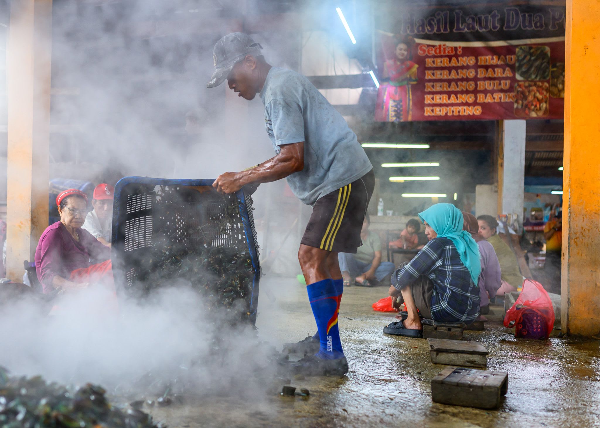A man tips a steaming basket of mussels in a market hall where women sit shelling.