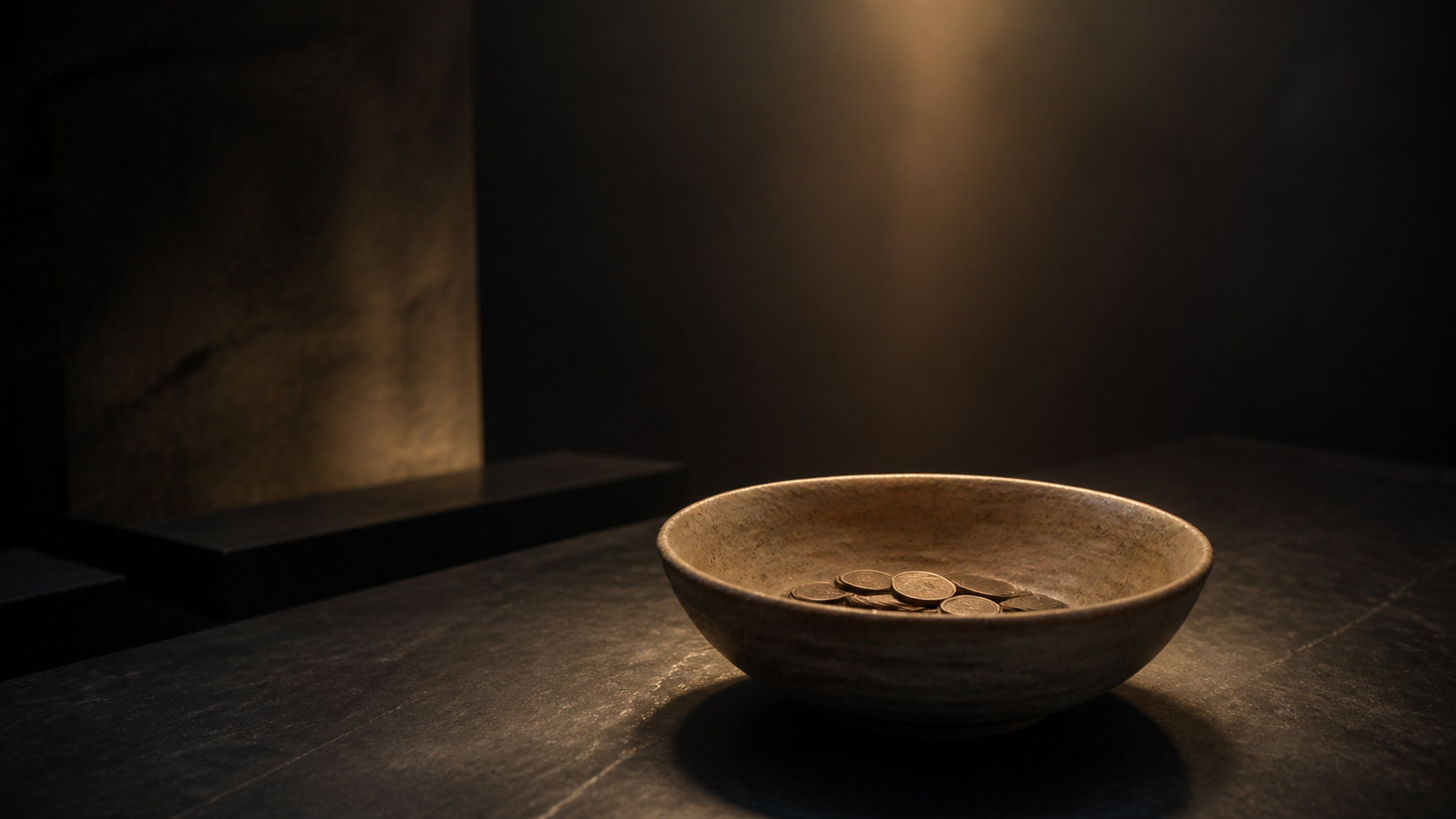 A modest ceramic bowl holding several coins beneath a calm beam of warm light in a dark studio