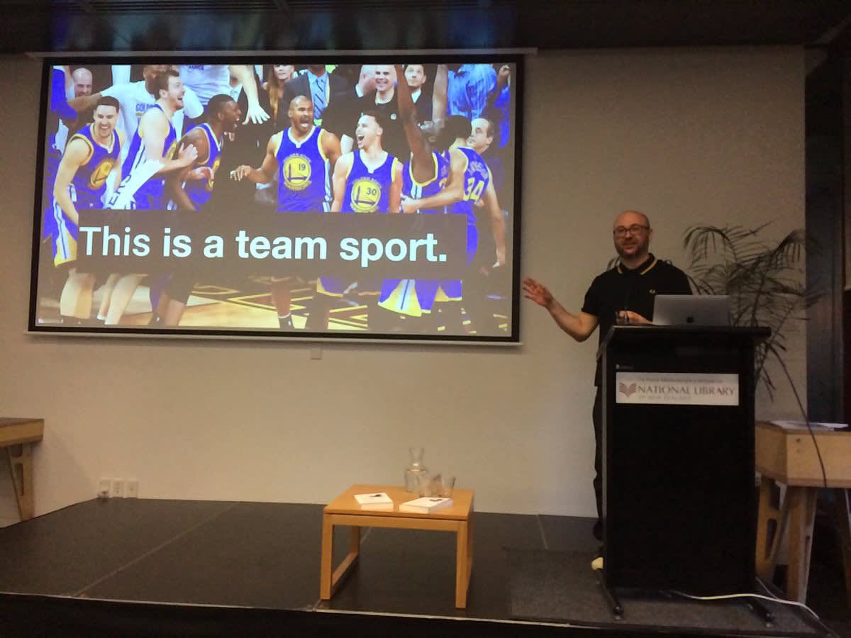 a guest presenter mid-speech standing in front of a projector screen with a slide that reads 'this is a team sport''