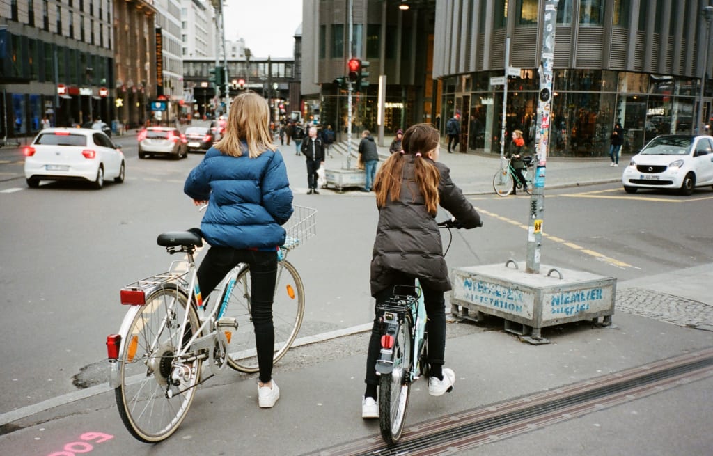 Two young girls on bikes, seen from behind, waiting for traffic at an intersection in a busy city