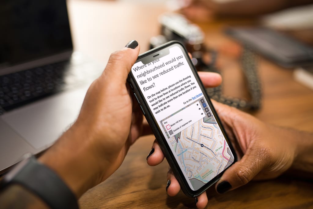 Woman's hands holding a smartphone displaying a geospatial question within Sutton's Neighbourhood Placemaking consultation. The map displays a user-drawn route.