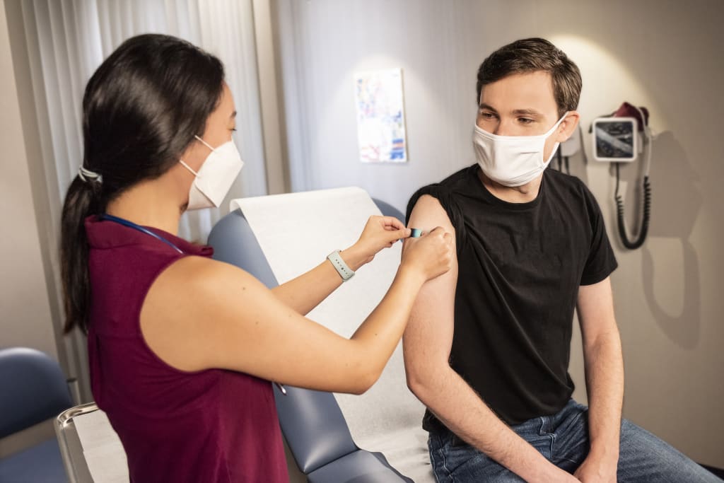 Female healthcare professional administering a vaccination to a young male. Both are wearing face masks