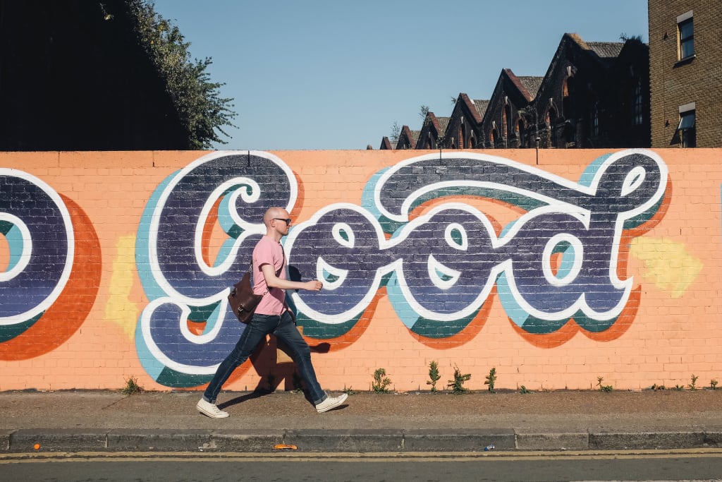 A photo of a man walking past some colourful street art which says the word good in big cursive letters.