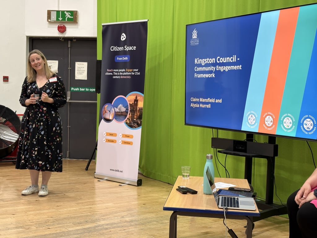 Claire Mansfield, a woman with long blonde hair wearing a dark patterned dress, stands looking at a large screen to her left. The screen displays a slide from her presentation about Kingston Council's Community Engagement Framework.