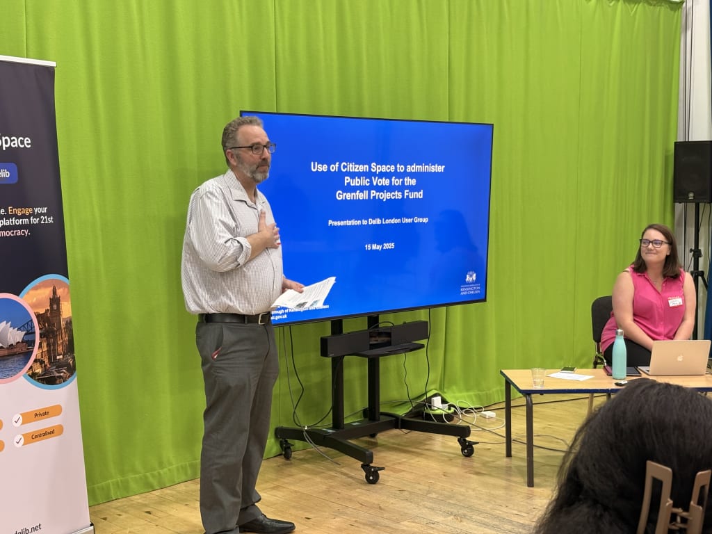 Gary Wilson, a man with salt-and-pepper coloured hair and beard wearing grey trousers and a pale shirt stands in front of a large screen to his left. The screen displays a slide from his presentation about Kensington and Chelsea Council's use of Citizen Space to administer the public vote fro the Grenfell Projects Fund.