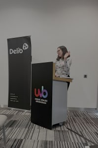 A young woman with shoulder-length dark brown hair wearing a glasses and a grey jumper (Ellen Taplin from Surrey County Council) stands behind a podium and addresses an audience (who are not pictured). She is gesturing casually with her left hand.