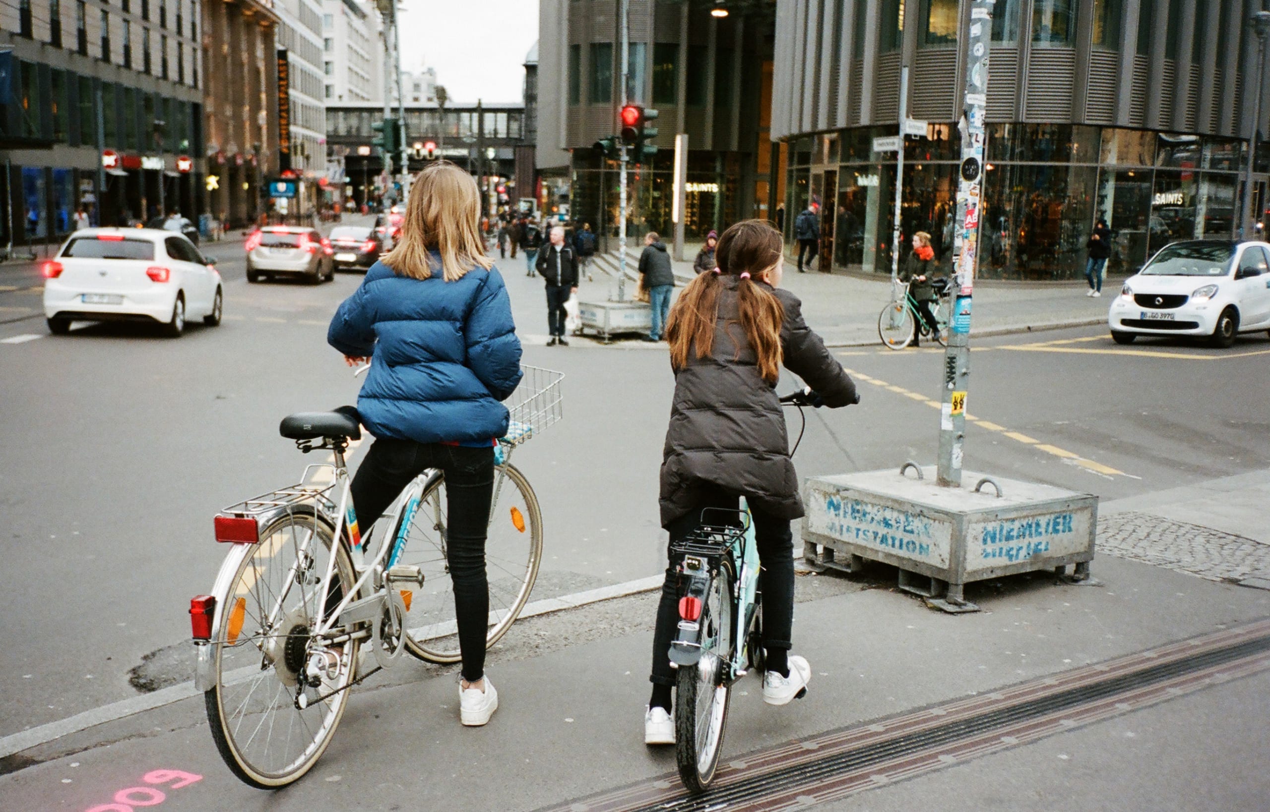 Two young girls on bikes, seen from behind, waiting for traffic at an intersection in a busy city