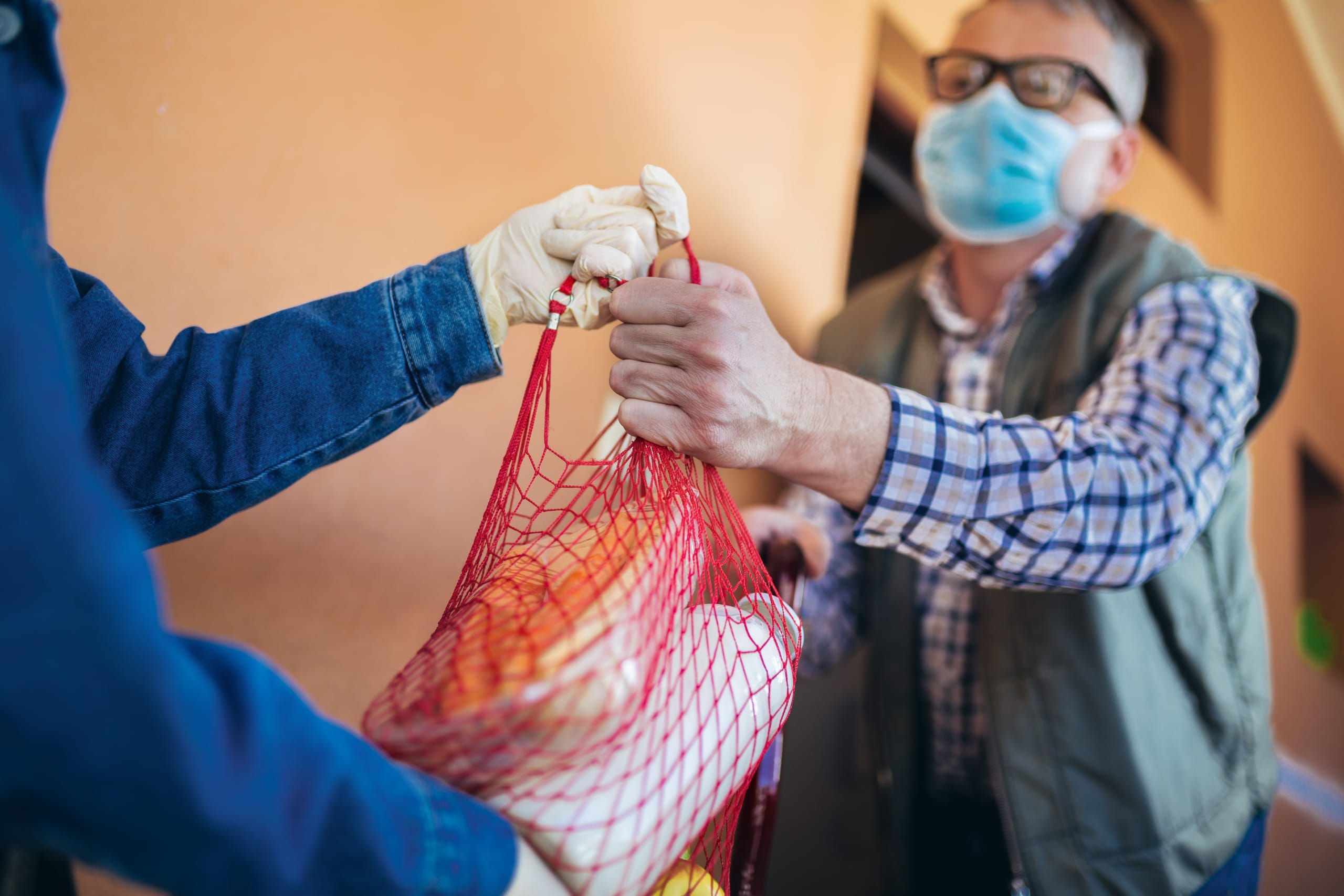 A volunteer with disposable gloves delivers groceries to an elderly man wearing a mask
