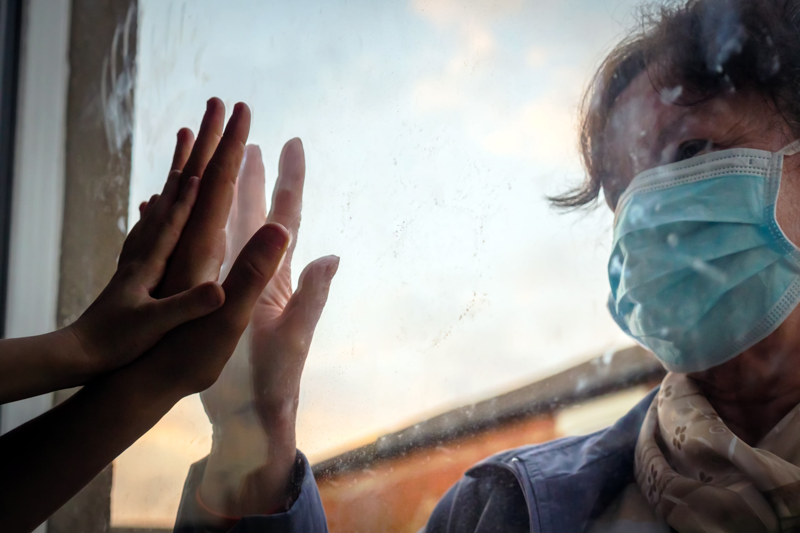 woman touches hands through glass with relative who is standing behind a window and wearing a mask