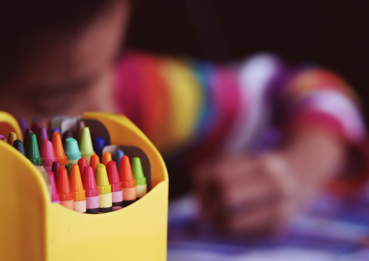 Close up of brightly coloured crayons whilst a child colours in soft focus in the background.