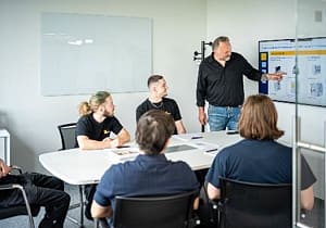 a man presenting machines on a screen with a group of people sitting at a table watching