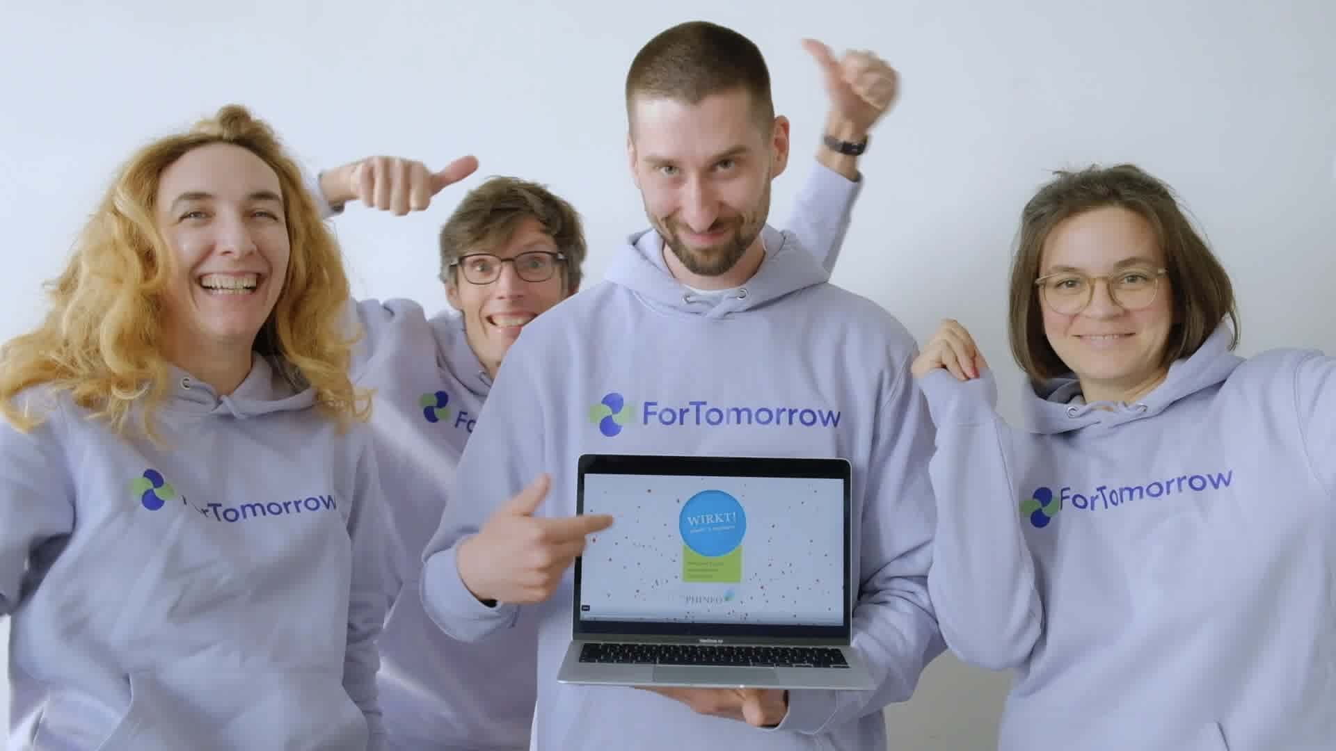 ForTomorrow-Team mit lila Hoodies steht vor der Kamera mit einem Laptop und jubelt. 