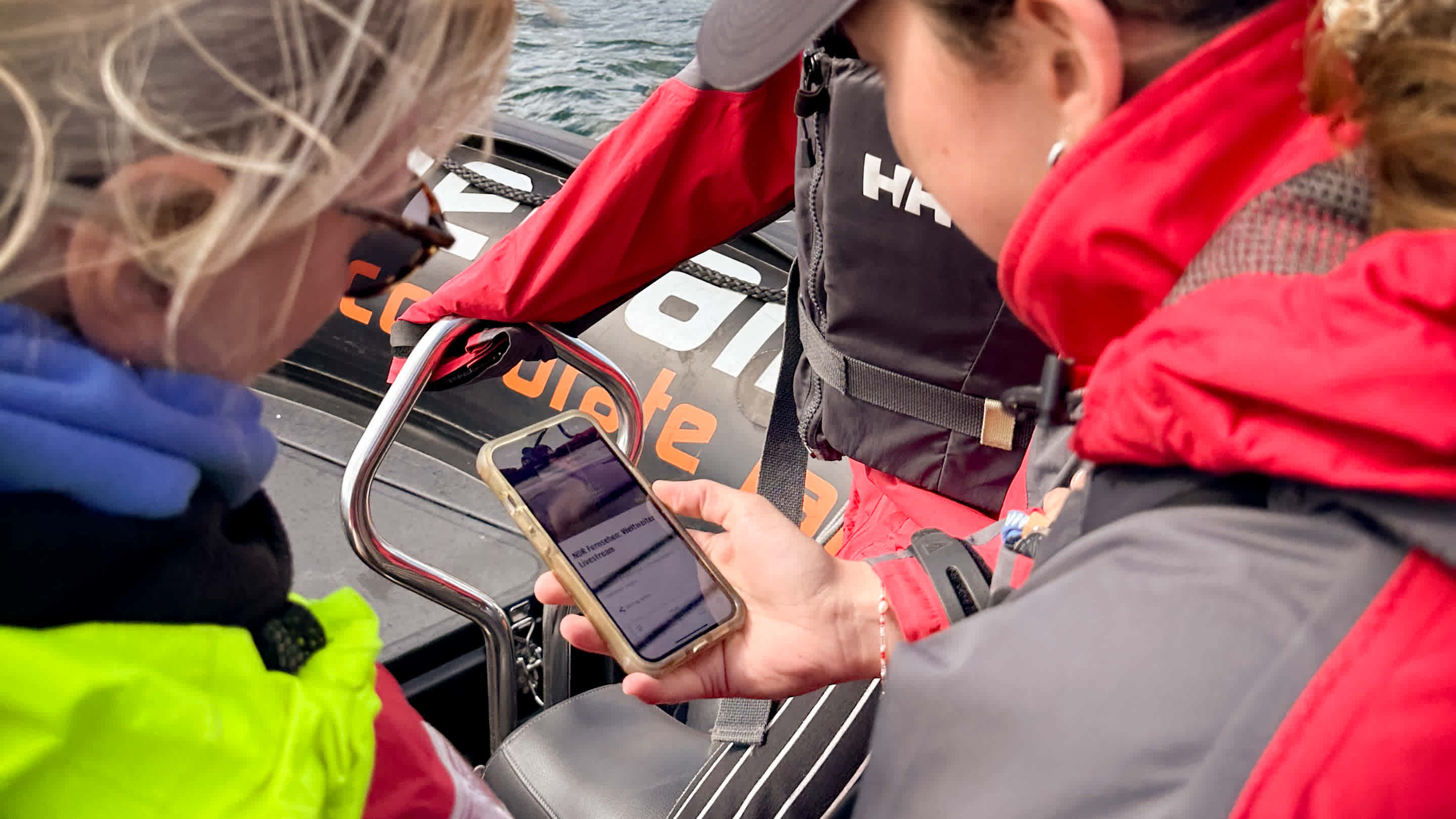 Auf einem Schnellboot steckt eine Gruppe Frauen die Köpfe zusammen, um auf einem Smartphone den Live-Stream vom Start des Rennens zu verfolgen.