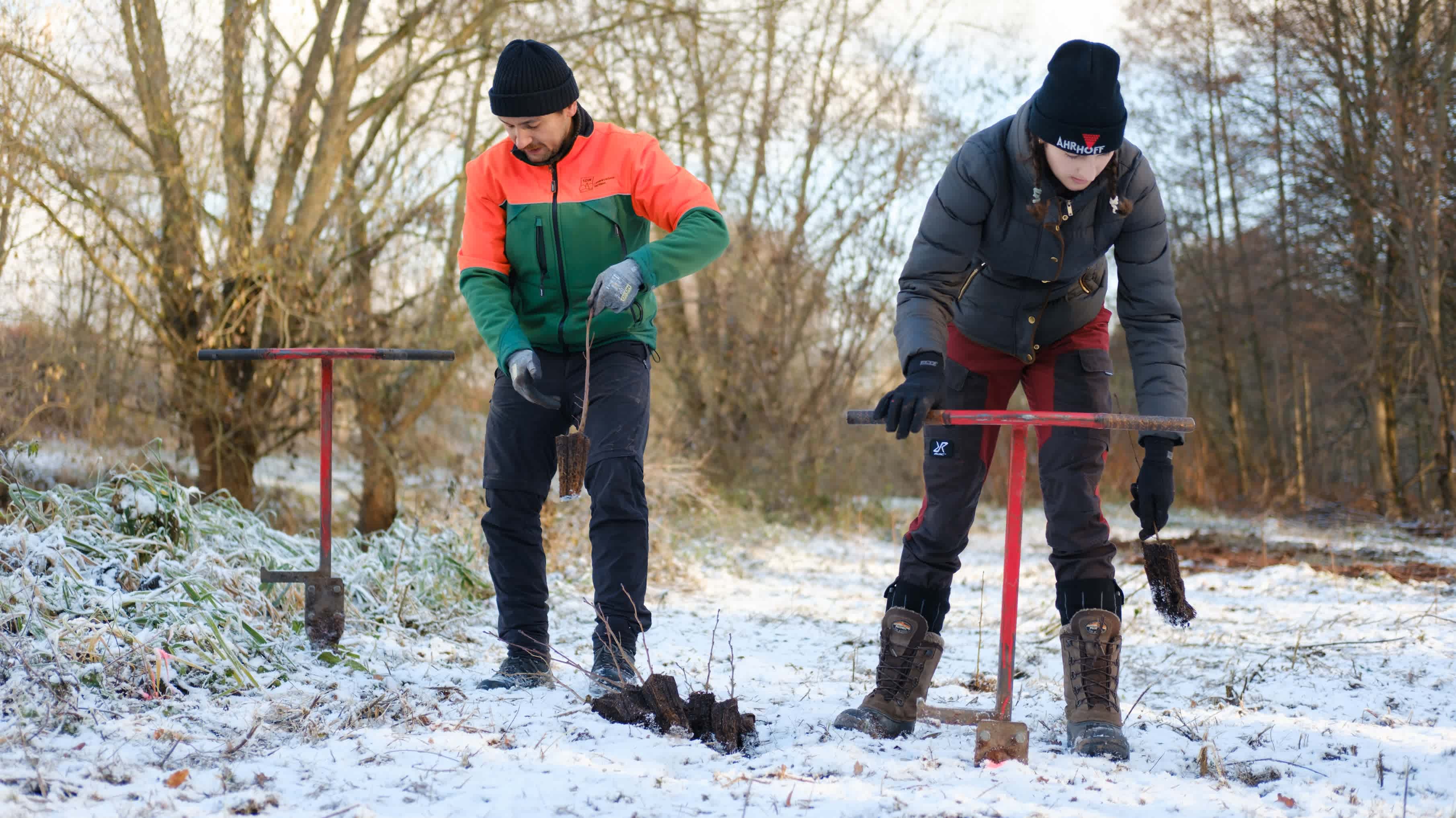 Two adults work the snow-covered meadow with planting tools.