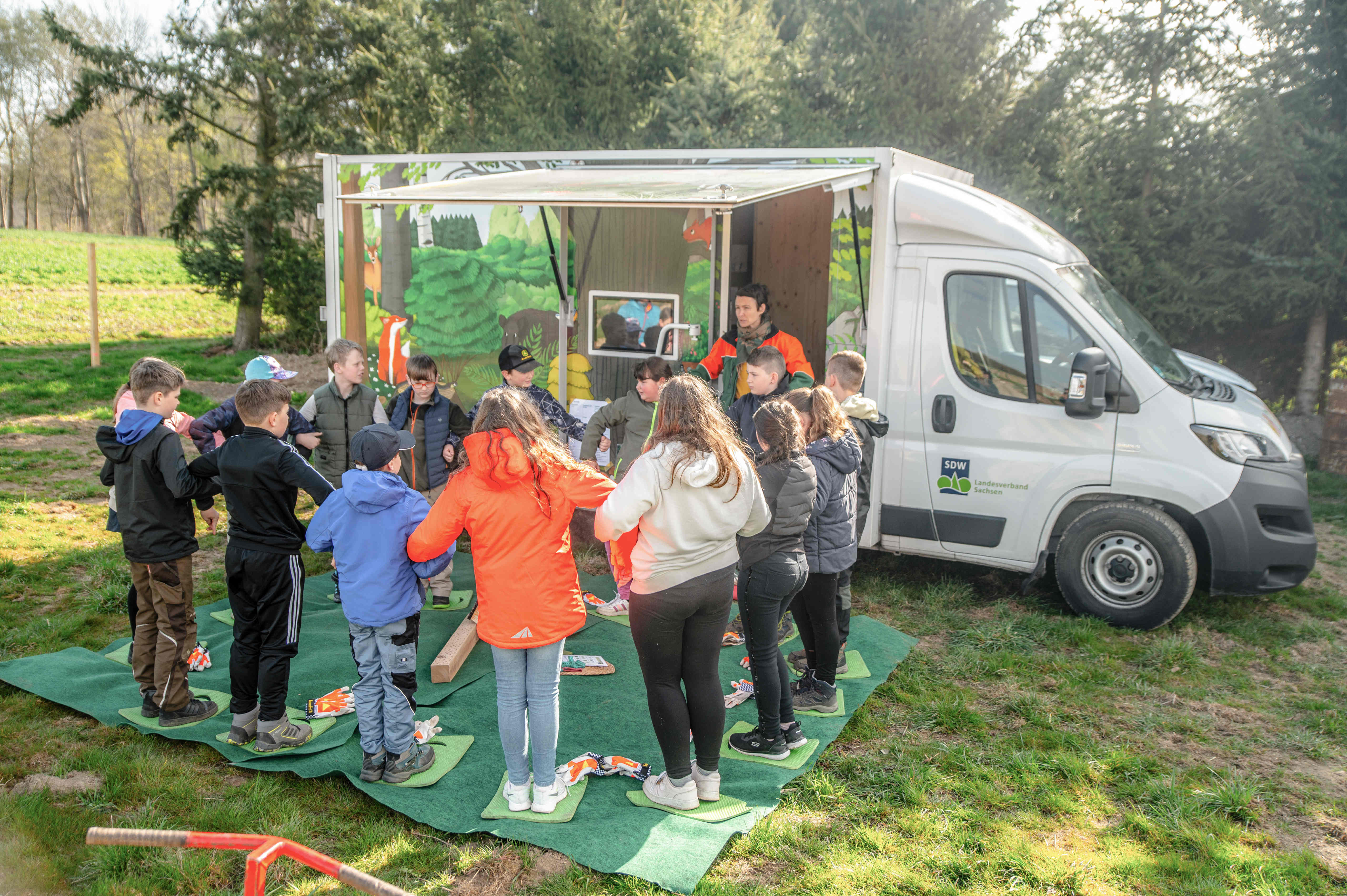 Die Schulklasse im Kreis mit Pädagogin vor einem bunten Bus 