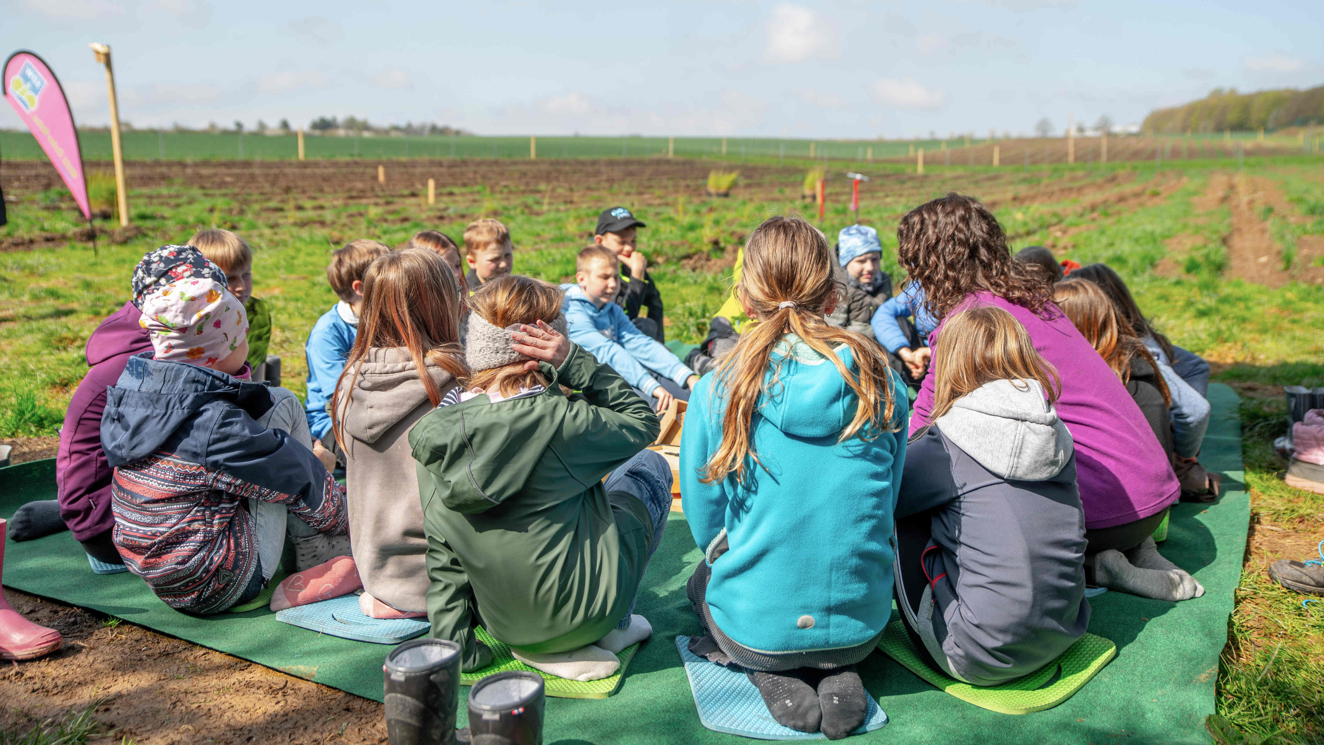 Die Pädagogin mit vielen Kindern, die auf einer großen Decke am Pflanzplatz sitzen