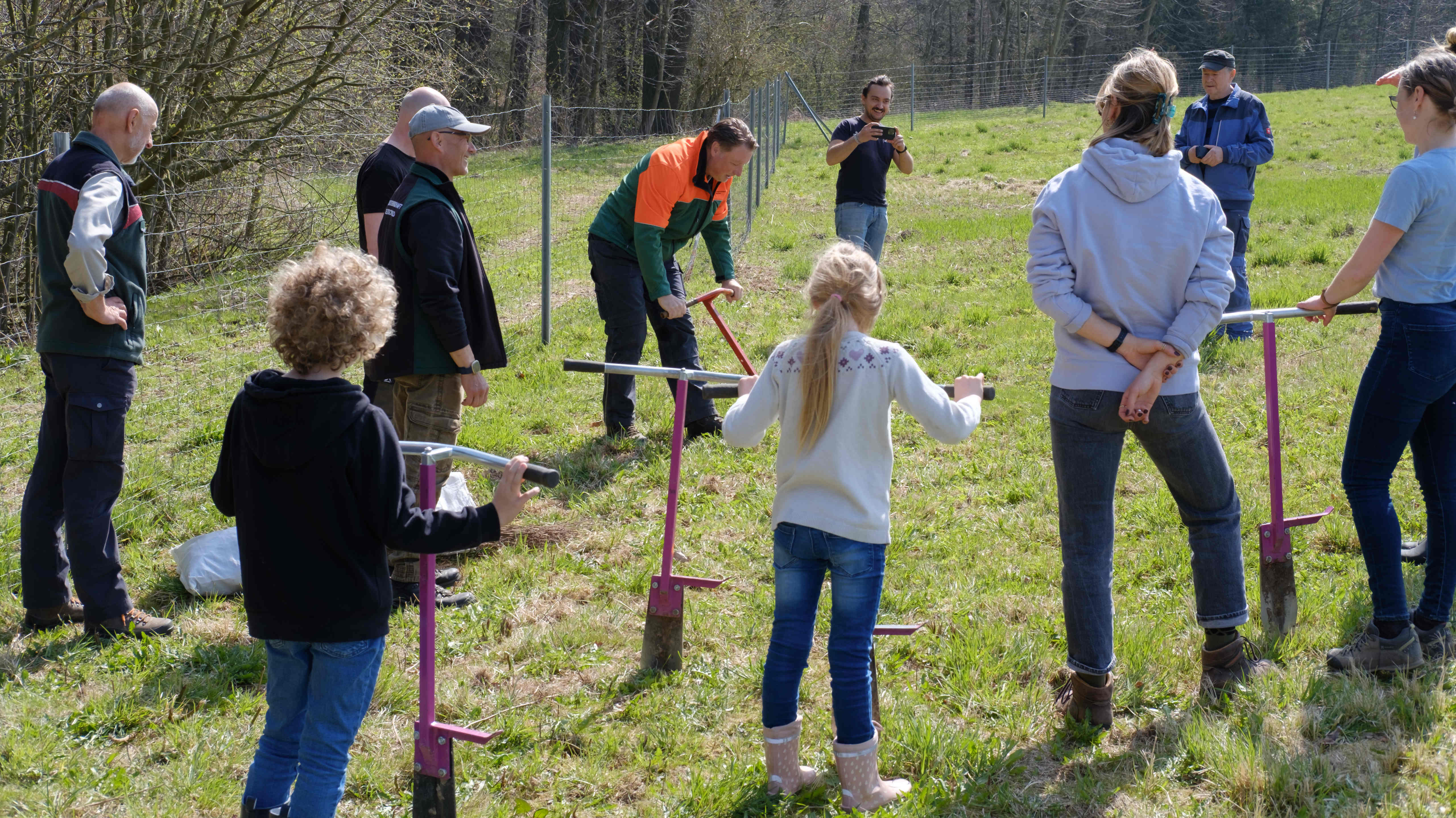 Gruppe Erwachsene und Kinder mit Baumpflanzspaten schauen auf einem Mann in Orange-Grüne SDW-Jacke.