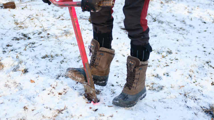 Heavy-booted feet push a planting spade into the snow-covered field