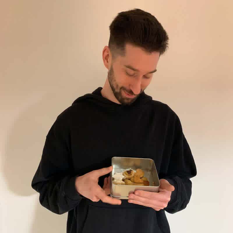 Chris von ForTomorrow steht vor einer weißen Wand, in seinen Händen befindet sich eine Keksdose, in die er lächelnd reinschaut. 