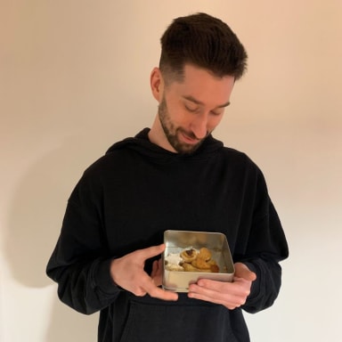Chris von ForTomorrow steht vor einer weißen Wand, in seinen Händen befindet sich eine Keksdose, in die er lächelnd reinschaut.
