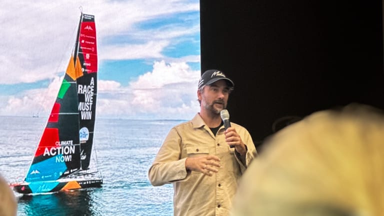 Boris Herrmann steht mit einem Mikrophon in der Hand vor einer Leinwand. Darauf zu sehen ist sein Boot mit dem Slogan Climate Action Now auf dem Segel.
