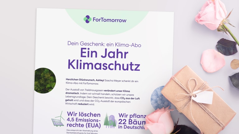 Geschenkentisch mit Rosen und einem ausgedruckten Zertifikat eines Klima-Abo-Geschenk. Der Text darauf ist: Dein Geschenk: ein Klima-Abo. Ein Jahr lang klimaneutral. Darunter Text, der zu klein ist zum Lesen. Sichtbar ist aber noch: 'Wir löschen 4,5 Emissionsrechte (EUA)' und 'Wir pflanzen 22 Bäume in Deutschland'