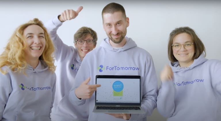 ForTomorrow-Team mit lila Hoodies steht vor der Kamera mit einem Laptop und jubelt.
