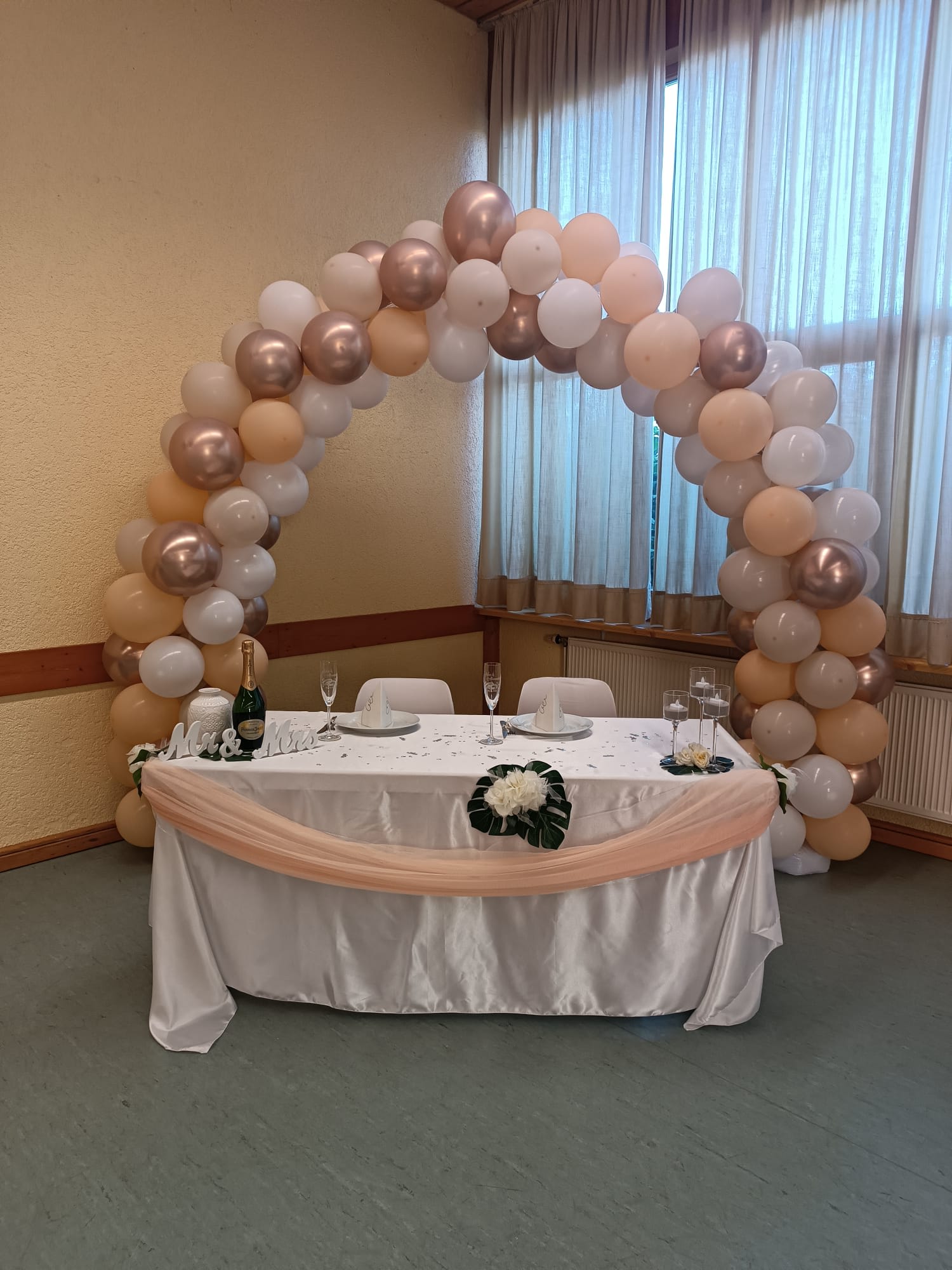 Dekorierter Tisch für das Brautpaar bei einer Hochzeit, dahinter ein Bogen aus bunten Luftballons