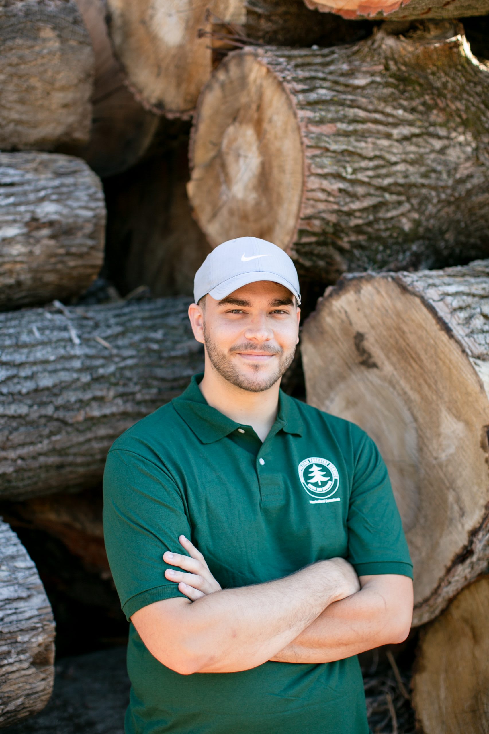 Man with arms folded wearing a tan hat and dark green shirt standing in front of tree logs.