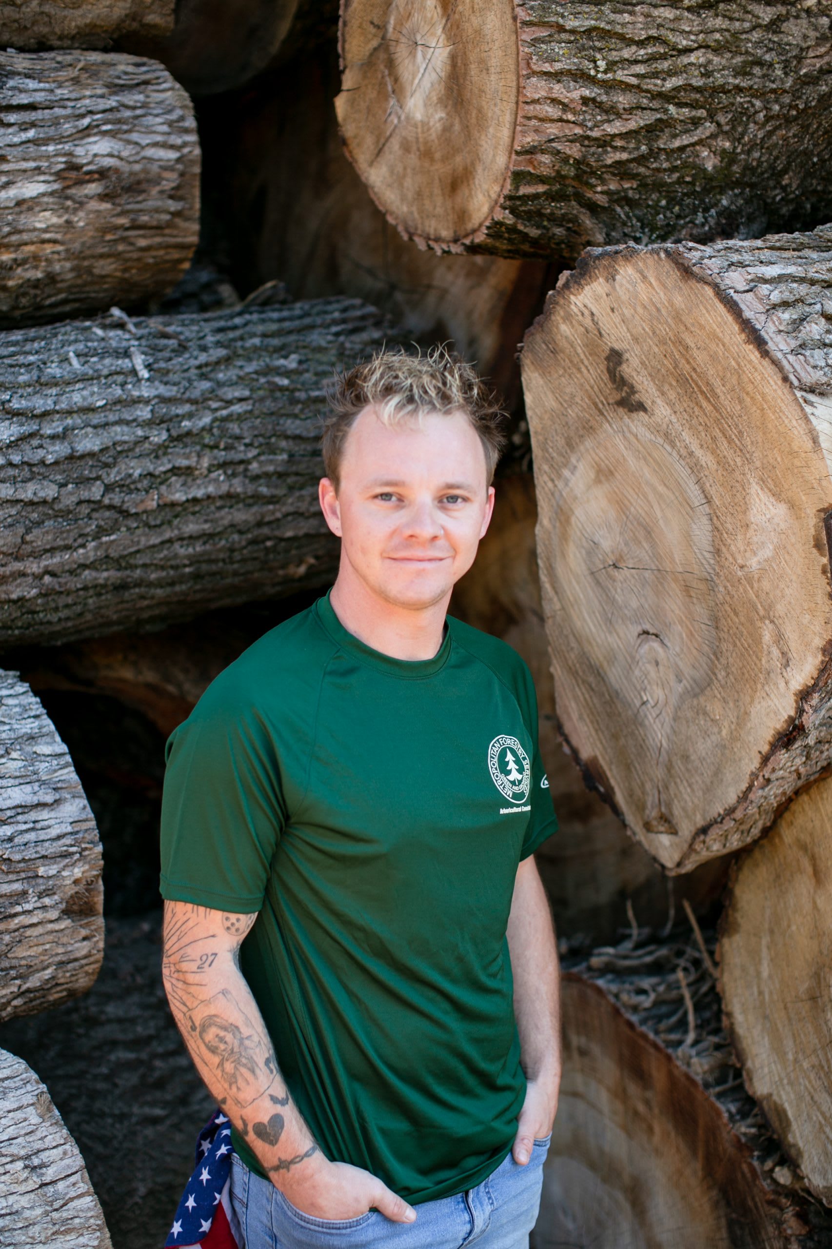 Man wearing a dark green shirt with his hands in his pockets standing in front of tree logs.