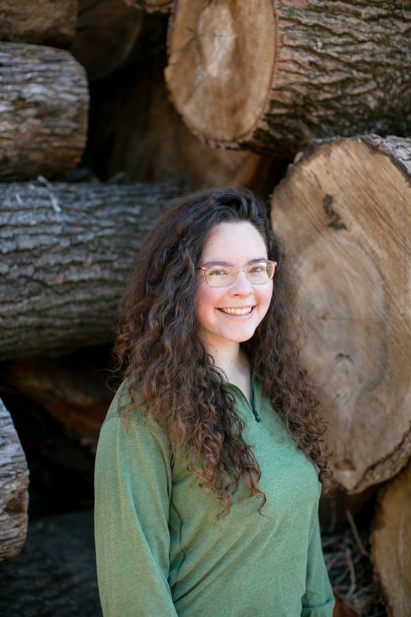 Woman with brown hair wearing light green shirt standing in front of tree logs.