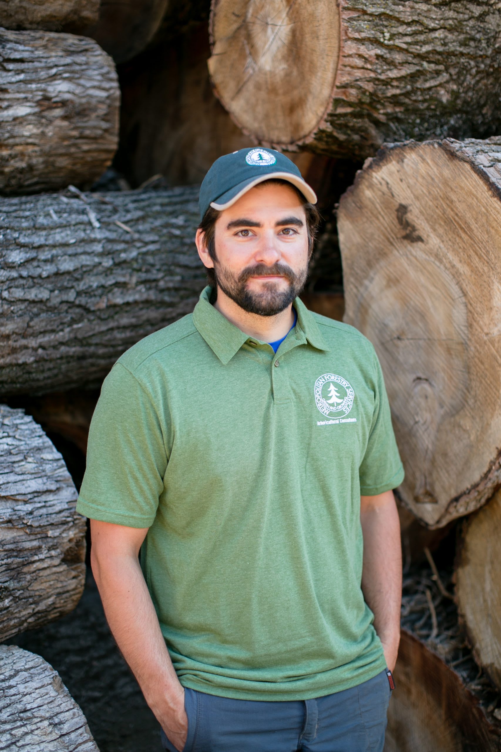 Man wearing a hat and a light green shirt with his hands in his pockets standing in front of tree logs.