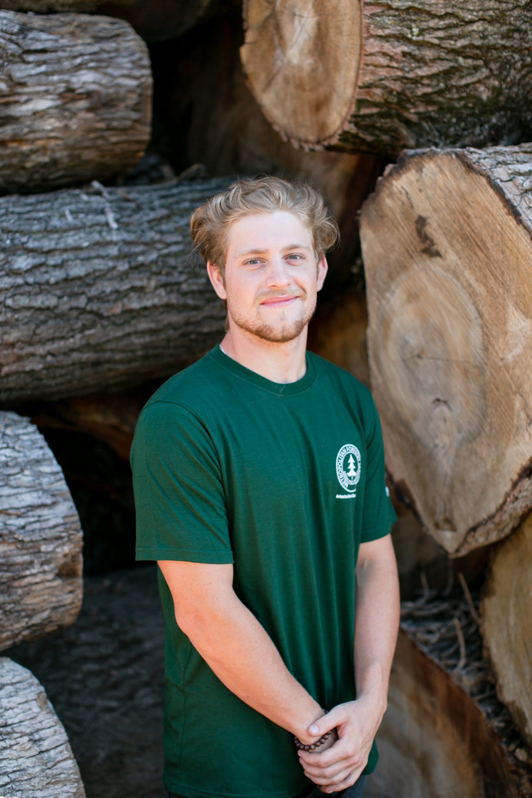 Man with hands clasped in front of him wearing a dark green shirt standing in front of tree logs