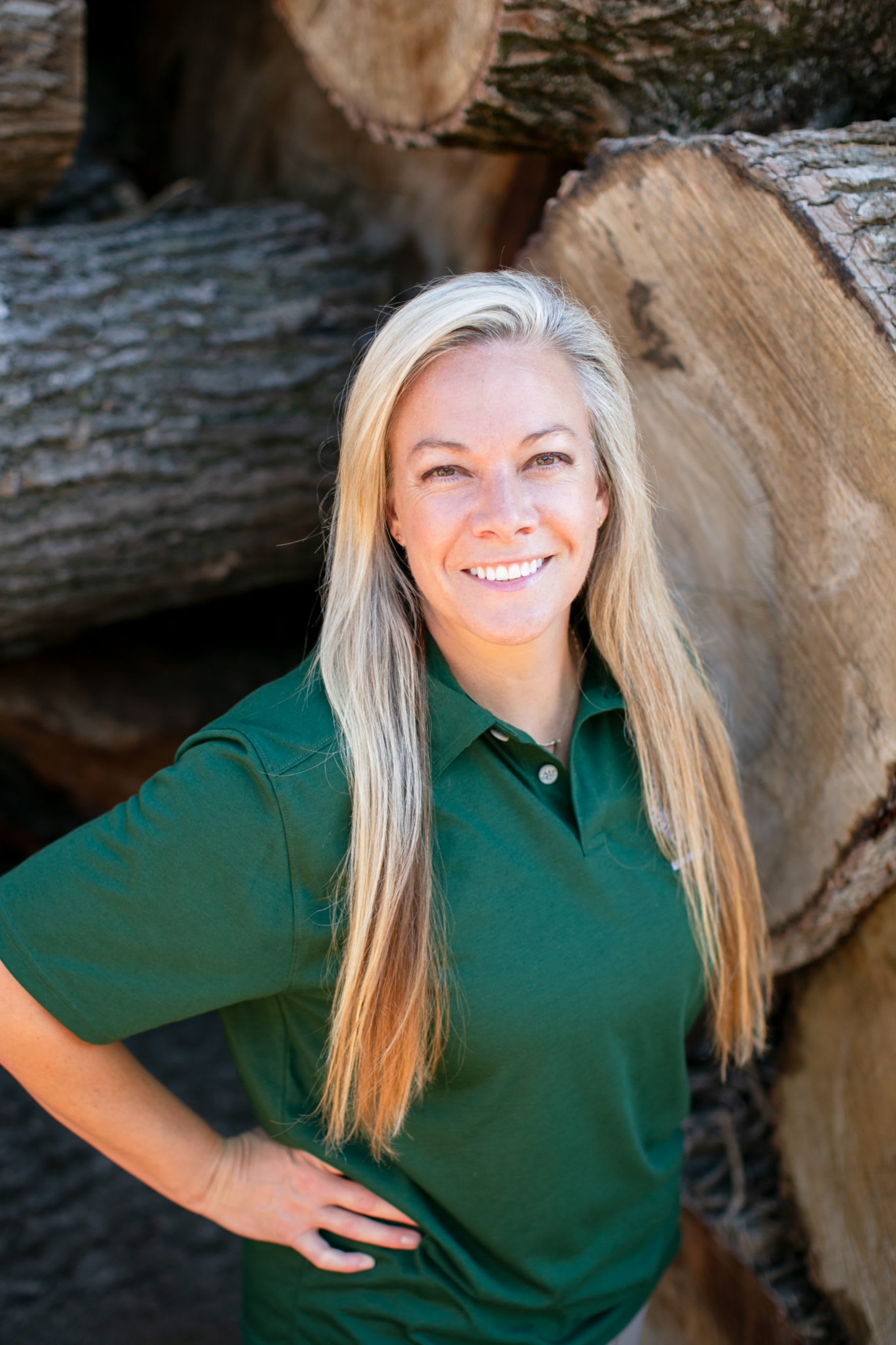 Woman with blond hair wearing a dark green shirt standing in front of tree logs.